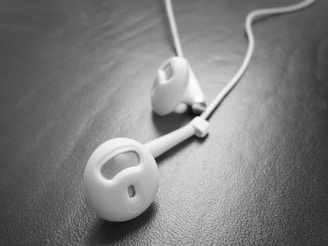 A close-up view of a pair of white earbud headphones resting on a textured, dark surface. The earbuds have a sleek and minimalist design with slight reflections on the surface, suggesting a smooth finish.