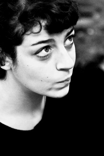 A black and white portrait of a person with medium-length curly hair, looking upwards. The focus is on the person's expressive eyes and contemplative expression. The background is blurred, drawing attention to the subject's face.