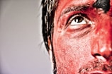 Close-up of a person's face with intense red makeup, capturing a detailed texture of the skin and hair. The person's eye looks upward, and the lighting highlights the contrast between the natural skin tone and the applied makeup.