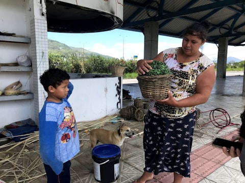 A woman holds a basket with green contents while a young boy stands nearby with his hand on his head. They are under a shelter with a tiled floor, and a dog is also present. Various materials and tools are scattered around, suggesting a crafting or workshop area. In the background, there is lush greenery and a hill, indicating an outdoor environment.