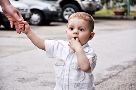 A toddler wearing a white shirt is walking outdoors while holding an adult's hand. The child has a curious expression, with one hand in their mouth. The background features parked cars and some greenery.