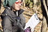 A person wearing a green knitted hat and red glasses is holding a clipboard with papers while standing in a forest area. They are dressed in a black jacket, and the background consists of trees with light filtering through the leaves.