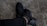 A pair of black athletic shoes are being worn by a person whose legs are crossed. The shoes are on a rough, grey textured surface, and the person is wearing black pants.