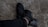 A pair of black athletic shoes are being worn by a person whose legs are crossed. The shoes are on a rough, grey textured surface, and the person is wearing black pants.