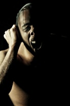A person is captured in low-key lighting, emphasizing stark contrasts and shadows. The face is partially visible, with an expression of intense emotion or anguish. The lighting highlights the contours and details of the muscles and features. The surrounding darkness adds a dramatic and moody effect.