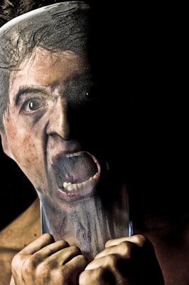 A person is seen with their face pressed against a translucent material, distorting their features and creating an expression of anguish or frustration. The image is dark, with a strong contrast between light and shadow on the face, emphasizing the emotional intensity.