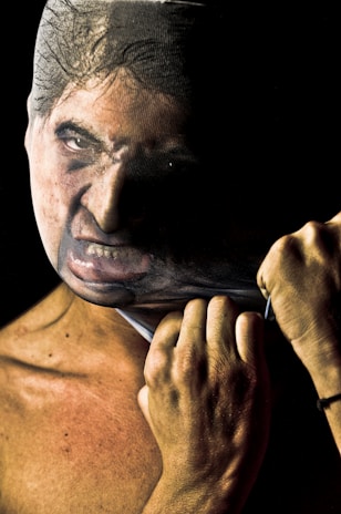 A person pulling a tight, face-printed stocking over their head, creating a distorted face with an intense expression. The lighting emphasizes the texture and details of the skin and fabric.