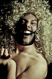 A person with curly blonde hair is laughing energetically. The face is partially covered with a dark mesh, adding a playful and mysterious element. The lighting creates a dramatic contrast, highlighting the facial expression and hair.