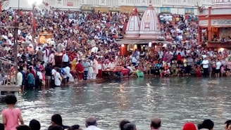 A large crowd gathers along the banks of a river, participating in a religious or cultural event. Many people are dressed in traditional attire. Temples and shrines can be seen in the background, and some individuals are near the water, possibly for ritual purposes.