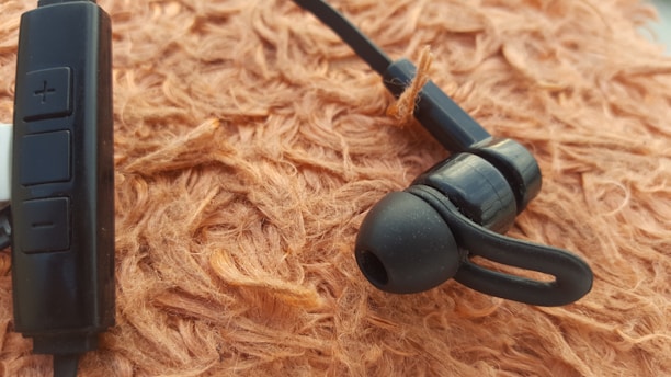 A close-up view of a black earphone resting on a textured, brownish-orange surface. The earphone features a small loop for securing it in the ear and a control module with buttons for adjusting audio settings.