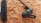 A close-up view of a black earphone resting on a textured, brownish-orange surface. The earphone features a small loop for securing it in the ear and a control module with buttons for adjusting audio settings.
