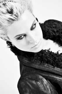 A monochrome portrait of a person with light-colored, voluminous hair, wearing dark eye makeup and giving an intense gaze. The person is dressed in a dark, textured jacket with a fluffy collar, and the image has a high-contrast aesthetic.