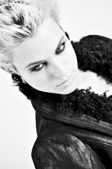 A monochrome portrait of a person with light-colored, voluminous hair, wearing dark eye makeup and giving an intense gaze. The person is dressed in a dark, textured jacket with a fluffy collar, and the image has a high-contrast aesthetic.