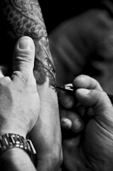 A close-up view of a tattoo being applied to someone's arm by a tattoo artist. The artist's hand is steady as they work on the intricate details of the design. The scene captures the intensity and focus required in tattooing.