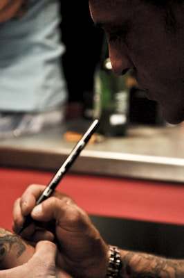 A tattoo artist is focused on creating a tattoo on a person's arm. The close-up view captures the precision of the tattooing process. The artist's hand is steady, holding a thin instrument, while the tattoo being worked on is partially visible. The background is blurred, emphasizing the concentration of the work.