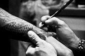 A person is drawing on a tattooed arm with a pen. The tattoo features intricate designs, possibly of a snake or dragon. The hands are focused on the detailing, suggesting precision. The monochromatic tone gives it an artistic and serious atmosphere.