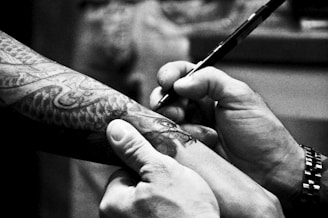 A person is drawing on a tattooed arm with a pen. The tattoo features intricate designs, possibly of a snake or dragon. The hands are focused on the detailing, suggesting precision. The monochromatic tone gives it an artistic and serious atmosphere.