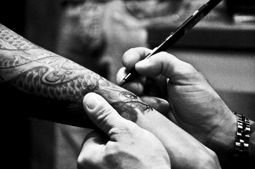 A person is drawing on a tattooed arm with a pen. The tattoo features intricate designs, possibly of a snake or dragon. The hands are focused on the detailing, suggesting precision. The monochromatic tone gives it an artistic and serious atmosphere.