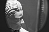 A black and white image of a sculpture depicting the profile of a serene and expressionless human face. The artwork features detailed, smooth facial contours and stylized hair arranged in sleek parallel lines. The softly illuminated figure is captured within an oval or circular frame, creating a focus on the sculpture's profile.