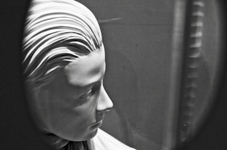 A black and white image of a sculpture depicting the profile of a serene and expressionless human face. The artwork features detailed, smooth facial contours and stylized hair arranged in sleek parallel lines. The softly illuminated figure is captured within an oval or circular frame, creating a focus on the sculpture's profile.