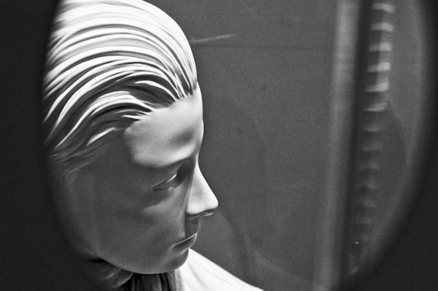 A black and white image of a sculpture depicting the profile of a serene and expressionless human face. The artwork features detailed, smooth facial contours and stylized hair arranged in sleek parallel lines. The softly illuminated figure is captured within an oval or circular frame, creating a focus on the sculpture's profile.
