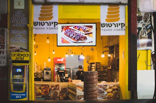 A brightly lit bakery shop with a yellow interior, displaying a variety of baked goods and a digital screen showcasing pastries. The counter is adorned with coffee machines and snack items. The store sign includes text in Hebrew and English, indicating available items such as birthday cake, babka, and coffee. An ATM is located on the left side.