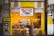 A brightly lit bakery shop with a yellow interior, displaying a variety of baked goods and a digital screen showcasing pastries. The counter is adorned with coffee machines and snack items. The store sign includes text in Hebrew and English, indicating available items such as birthday cake, babka, and coffee. An ATM is located on the left side.
