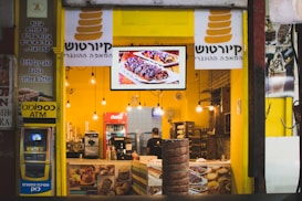 A brightly lit bakery shop with a yellow interior, displaying a variety of baked goods and a digital screen showcasing pastries. The counter is adorned with coffee machines and snack items. The store sign includes text in Hebrew and English, indicating available items such as birthday cake, babka, and coffee. An ATM is located on the left side.