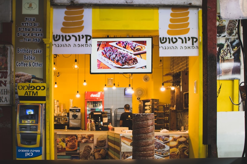 A brightly lit bakery shop with a yellow interior, displaying a variety of baked goods and a digital screen showcasing pastries. The counter is adorned with coffee machines and snack items. The store sign includes text in Hebrew and English, indicating available items such as birthday cake, babka, and coffee. An ATM is located on the left side.