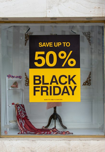 A yellow and black poster in a shop window reading 50% Black Friday, now it's time to save big, with a shoe on a shelf behind it
