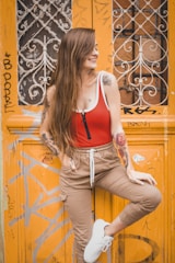 A young woman with long hair, wearing a red and white top and beige pants, poses confidently in front of an ornate, graffiti-covered orange door. She has tattoos on her arms and is wearing white sneakers. The door features intricate metalwork and urban graffiti, adding an artistic backdrop.