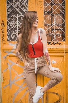 A young woman with long hair, wearing a red and white top and beige pants, poses confidently in front of an ornate, graffiti-covered orange door. She has tattoos on her arms and is wearing white sneakers. The door features intricate metalwork and urban graffiti, adding an artistic backdrop.
