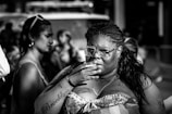 A black and white image featuring a person with braided hair and glasses, holding a hand to their mouth. The person has visible tattoos and is wearing a strapless top. In the background, blurred figures and urban elements are present, suggesting a street or public setting.