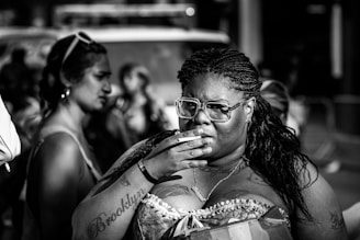 A black and white image featuring a person with braided hair and glasses, holding a hand to their mouth. The person has visible tattoos and is wearing a strapless top. In the background, blurred figures and urban elements are present, suggesting a street or public setting.
