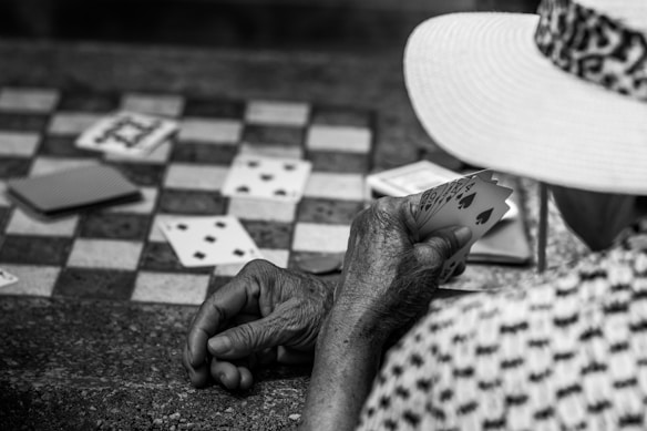 An elderly person wearing a patterned hat is playing cards on a stone or concrete surface with a checkerboard pattern. A few playing cards are scattered on the board.