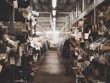A dimly lit warehouse aisle filled with rolls of fabric stored on metal shelves. Various tags and labels are attached to the fabric rolls, and the passageway appears to be narrow, lined with metal racks on both sides.