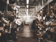 A dimly lit warehouse aisle filled with rolls of fabric stored on metal shelves. Various tags and labels are attached to the fabric rolls, and the passageway appears to be narrow, lined with metal racks on both sides.