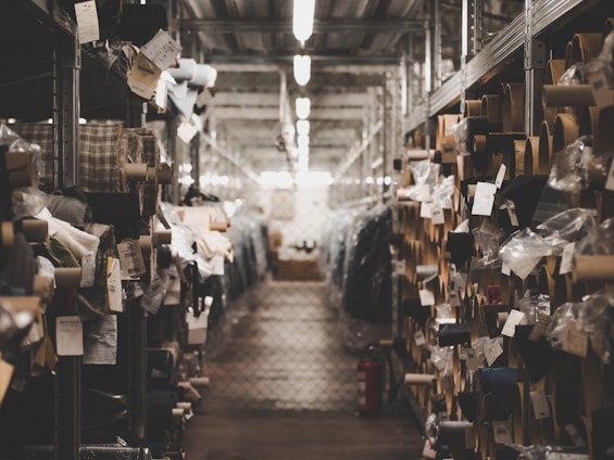 A dimly lit warehouse aisle filled with rolls of fabric stored on metal shelves. Various tags and labels are attached to the fabric rolls, and the passageway appears to be narrow, lined with metal racks on both sides.