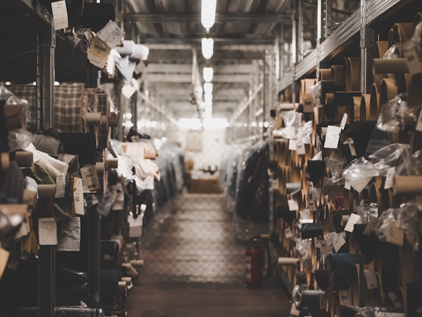 A dimly lit warehouse aisle filled with rolls of fabric stored on metal shelves. Various tags and labels are attached to the fabric rolls, and the passageway appears to be narrow, lined with metal racks on both sides.