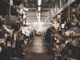 A dimly lit warehouse aisle filled with rolls of fabric stored on metal shelves. Various tags and labels are attached to the fabric rolls, and the passageway appears to be narrow, lined with metal racks on both sides.