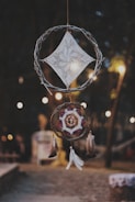 A dreamcatcher composed of two circular woven structures is hanging outdoors. The top circle features a knitted geometric pattern resembling a diamond shape, while the lower circle incorporates an intricate design with colorful strings and feathers attached at the bottom. The background is softly blurred, showing warm lights that hint at a cozy, evening setting.