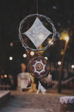 A dreamcatcher composed of two circular woven structures is hanging outdoors. The top circle features a knitted geometric pattern resembling a diamond shape, while the lower circle incorporates an intricate design with colorful strings and feathers attached at the bottom. The background is softly blurred, showing warm lights that hint at a cozy, evening setting.