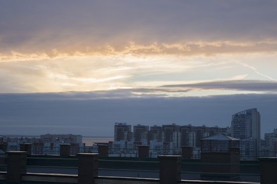 A cityscape at sunset with high-rise buildings silhouetted against a dramatic sky. The skyline features a variety of apartment and office buildings, with one distinctive curved structure on the right. The sky is layered with clouds, showing patches of golden light breaking through toward the horizon.