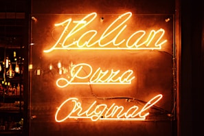 Neon sign with glowing orange letters reading 'Italian Pizza Original' against a dark background. The lighting creates a warm and inviting atmosphere, with visible reflections on the surface.