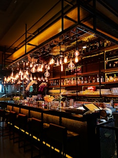 A modern bar with a sleek design, featuring a long counter with high chairs and shelves stocked with various bottles and glasses. Warm pendant lights hang overhead, casting a cozy glow. Decorative flowers and a few bar tools are placed on the counter.