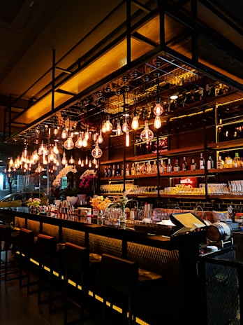 A modern bar with a sleek design, featuring a long counter with high chairs and shelves stocked with various bottles and glasses. Warm pendant lights hang overhead, casting a cozy glow. Decorative flowers and a few bar tools are placed on the counter.