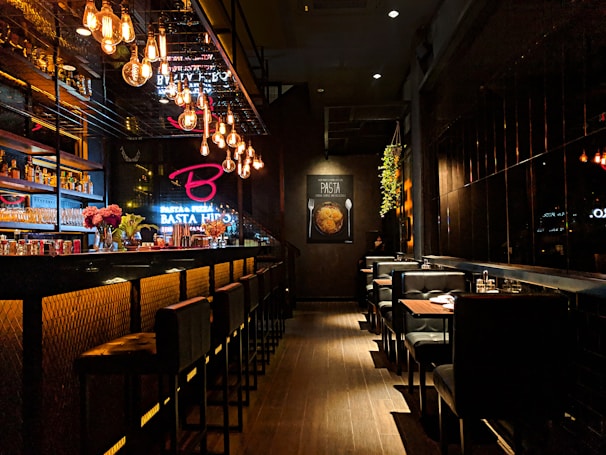 A dimly lit, stylish bar with modern decor, featuring a long counter on the left adorned with bottles and glassware. Hanging Edison bulb lights create a warm ambiance. The bar stools have a padded black design, while booths with neatly set tables line the right side. Wall art with pasta imagery adds a cozy touch, and small floral arrangements add vibrant color.
