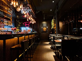 A dimly lit, stylish bar with modern decor, featuring a long counter on the left adorned with bottles and glassware. Hanging Edison bulb lights create a warm ambiance. The bar stools have a padded black design, while booths with neatly set tables line the right side. Wall art with pasta imagery adds a cozy touch, and small floral arrangements add vibrant color.