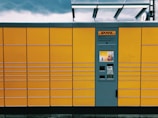 A yellow package locker system with a grey central panel featuring a screen and several compartments. The brand logo is visible above the panel. The structure has horizontal lines creating rectangular sections. The top of the locker has a transparent overhang, and the sky is overcast with dark clouds.