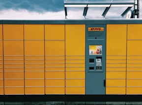 A yellow package locker system with a grey central panel featuring a screen and several compartments. The brand logo is visible above the panel. The structure has horizontal lines creating rectangular sections. The top of the locker has a transparent overhang, and the sky is overcast with dark clouds.